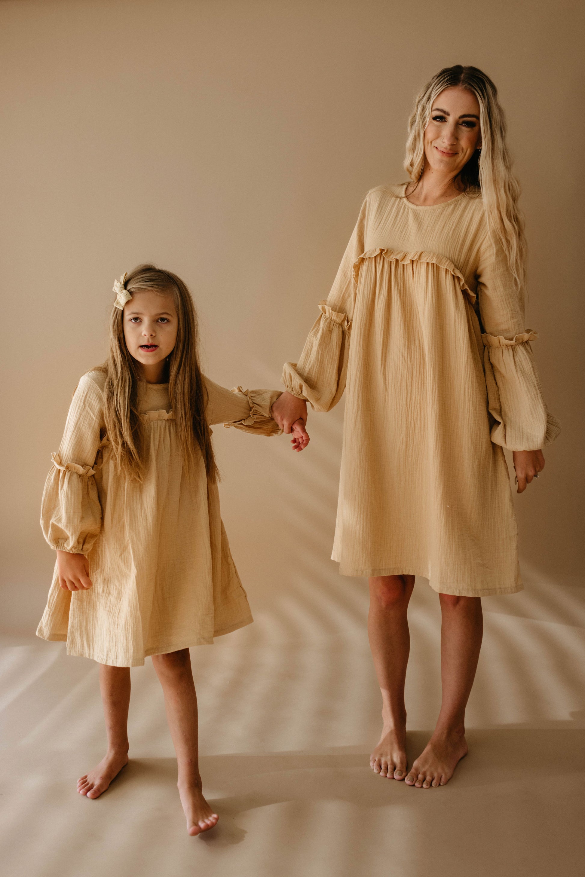 A woman and a young girl stand barefoot on a beige background, highlighting coordinated family outfits. They are both wearing "Golden Hour" light brown, long-sleeved dresses by forever french baby. The woman holds the girl's hand, and they both have long, wavy hair.