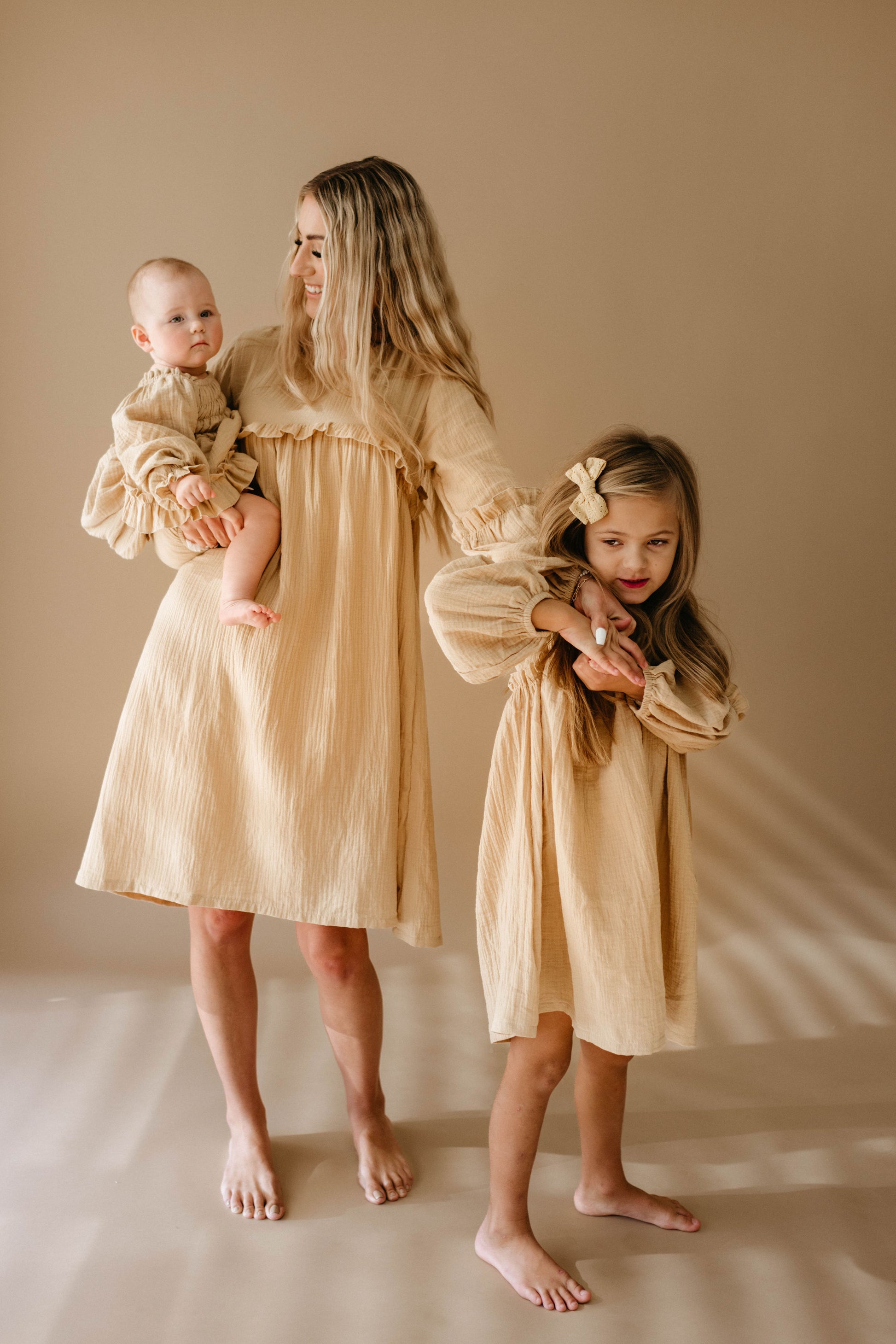 A woman holding a baby, dressed in the adorable "Golden Hour" Ruffle Romper from forever french baby, and standing beside a young girl, all barefoot in matching beige dresses with a golden hue. The young girl, cradling a small object, stands slightly ahead while the baby, snuggly wearing the romper available in sizes newborn to 24 months, rests on the woman's hip.