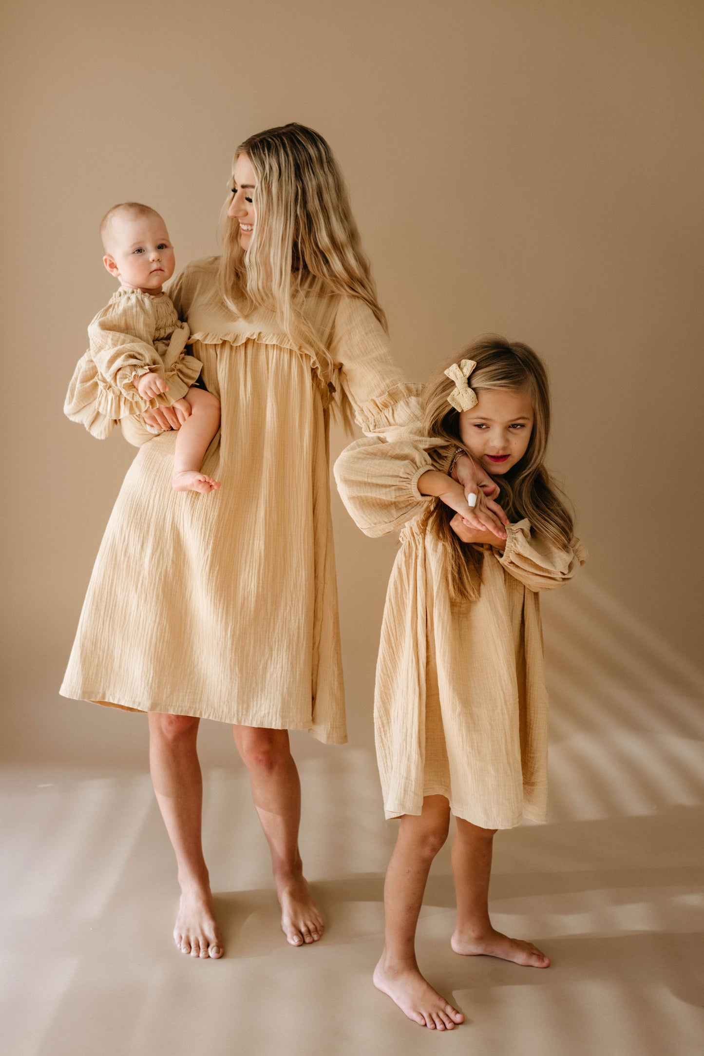 A woman holding a baby stands beside a young girl, all dressed in matching family outfits from the "Golden Hour" collection by forever french baby. Their light beige dresses with a subtle golden hue beautifully complement the warm, neutral background. They are barefoot on the floor, adding to the serene and harmonious atmosphere.