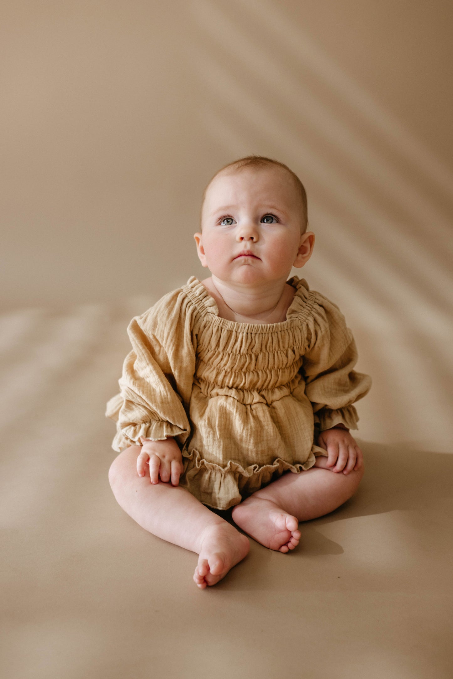 A baby sits barefoot on a brown background, dressed in the "Ruffle Romper | Golden Hour" by forever french baby. The baby gazes upward with a curious expression as soft, natural light casts gentle golden shadows, enhancing the serene scene. This outfit is available for sizes newborn to 24 months.