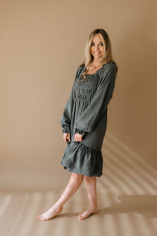 A woman with long blonde hair smiles barefoot on a beige floor, wearing the forever french baby Women's Dress | Meadow, a long-sleeved, knee-length dark gray dress, set against a soft tan background.