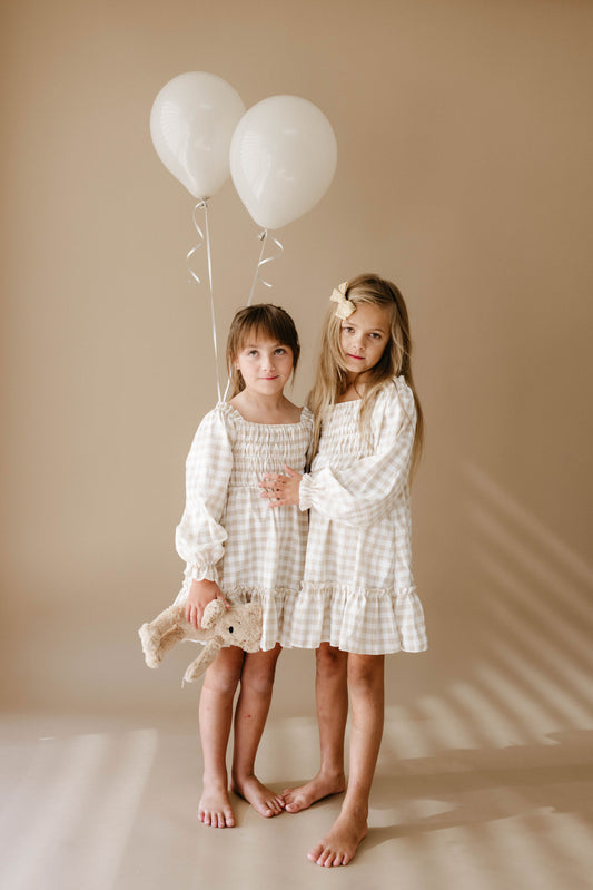 Two young girls stand barefoot on a neutral background, each wearing matching Child Dress | Gingham from forever french baby. One girl holds a teddy bear, while the other holds two white balloons. They both have long hair and stand close together, looking at the camera like models for these adorable dresses in baby sizes.