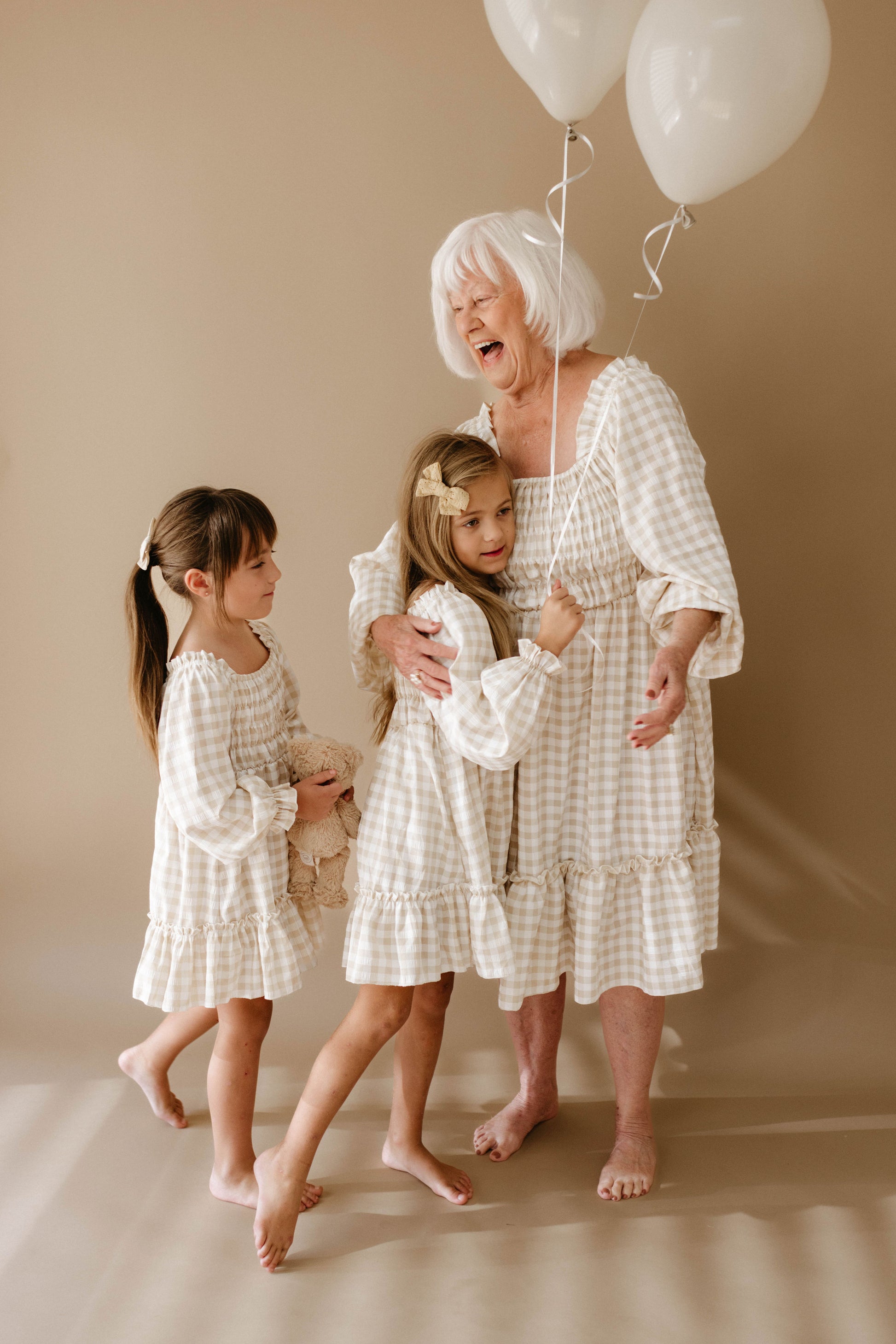 An elderly woman with white hair, clad in a checkered dress, holds two white balloons. She joyfully interacts with two young girls wearing dresses from the "Women's Dress | Gingham" collection by forever french baby, gleefully matching her style, in a warmly-lit room with a beige background.