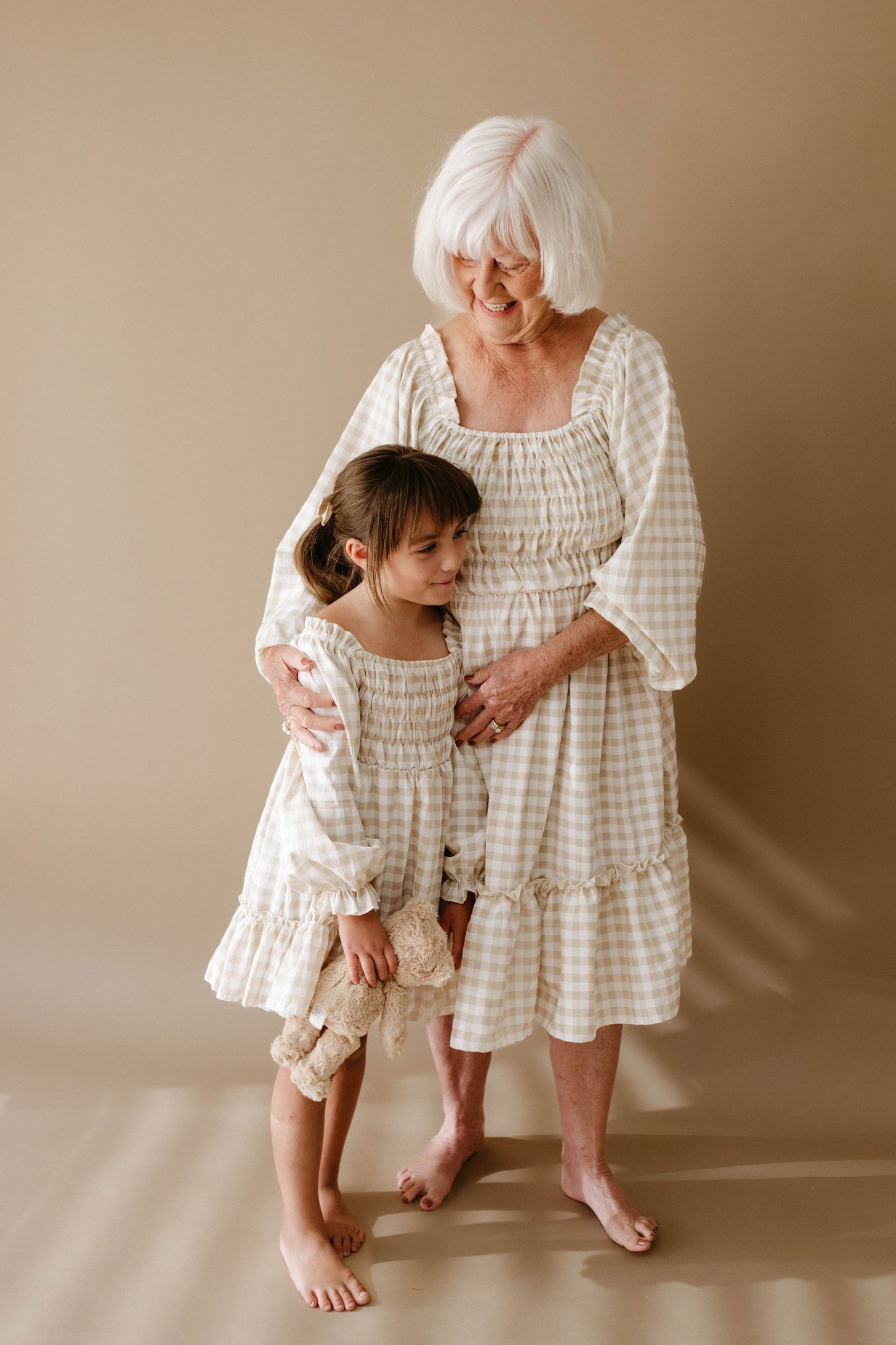 An elderly woman with white hair and a young girl stand together against a beige backdrop, both wearing matching forever french baby Women's Dress in gingham print. The woman smiles as she gently holds the girl, who is looking down while clutching her teddy bear.