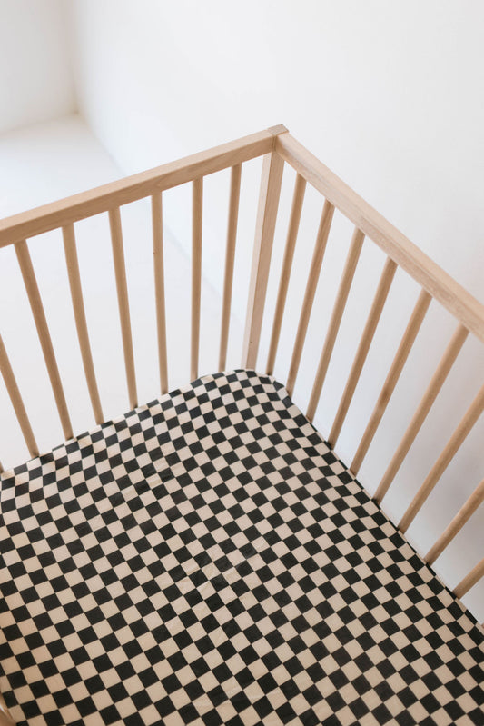 A wooden crib with vertical slats is pictured. The Muslin Crib Sheet by forever french baby, featuring a black and ivory checkerboard pattern, covers the mattress. The background is white and minimalistic, with natural light illuminating the scene.