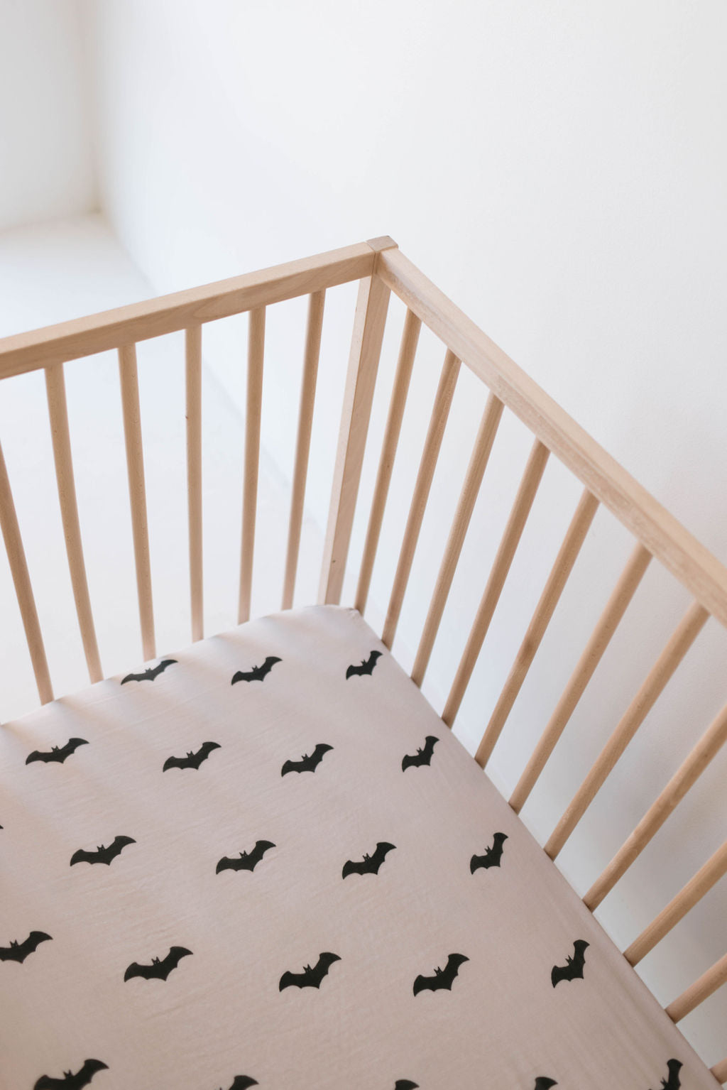 A wooden baby crib with vertical slats is pictured with the forever french baby's Muslin Crib Sheet | It's Bats!, decorated with small black bat designs. This charming nursery setup, against a plain white wall, creates a minimalist aesthetic.