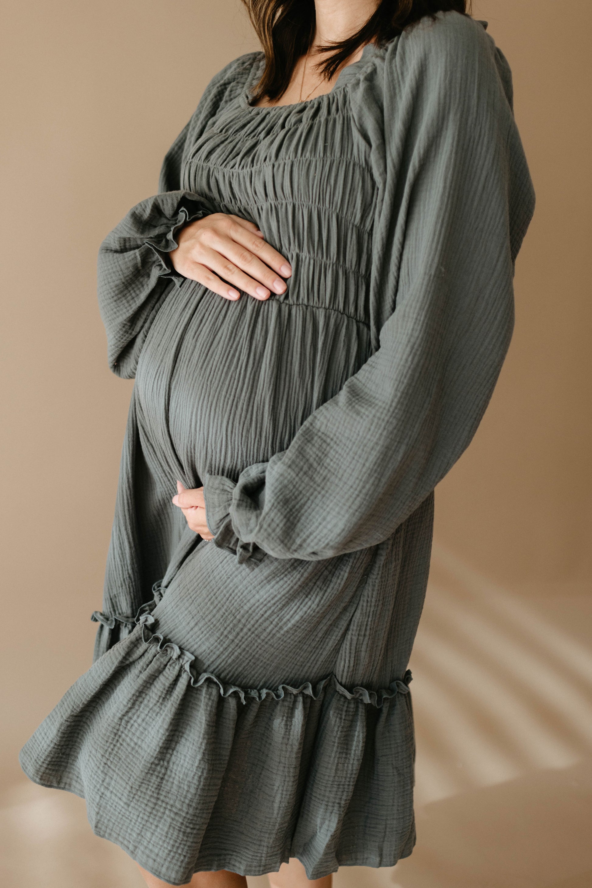 A pregnant woman, face unseen, gently cradles her belly in a loose, textured Women's Dress | Meadow by forever french baby against a soft beige background.