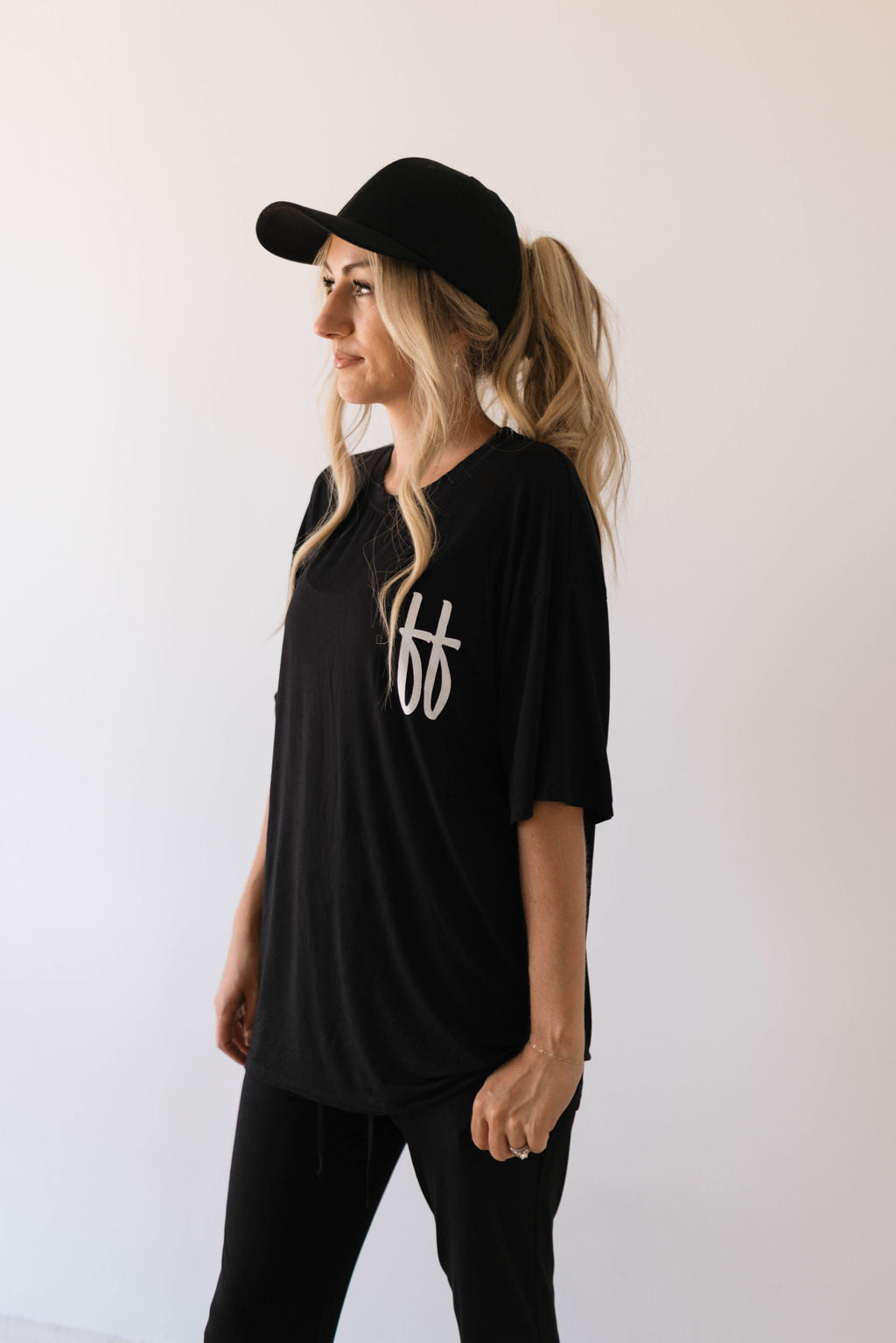 A person with long blonde hair tied in a ponytail, donning a black baseball cap and the "Short Sleeve Women's Bamboo Lounge Set | Black ff Signature" by forever french baby, stands against a plain white background, looking to the left. They are also wearing black pants.