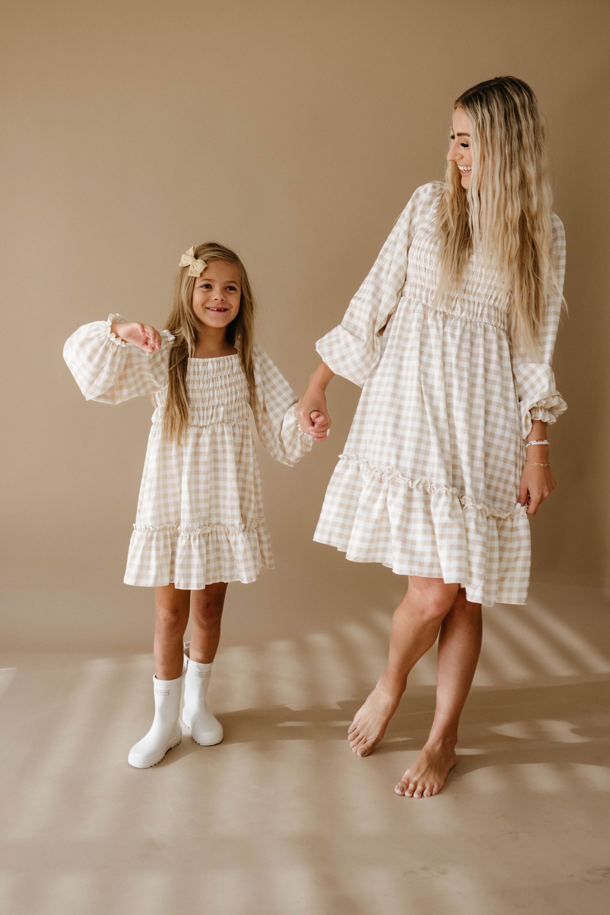 A woman and a young girl with long blonde hair hold hands and exchange smiles as they wear matching "Women's Dress | Gingham" by forever french baby. The young girl accessorizes with white boots, and the neutral beige background beautifully complements their coordinated outfits.