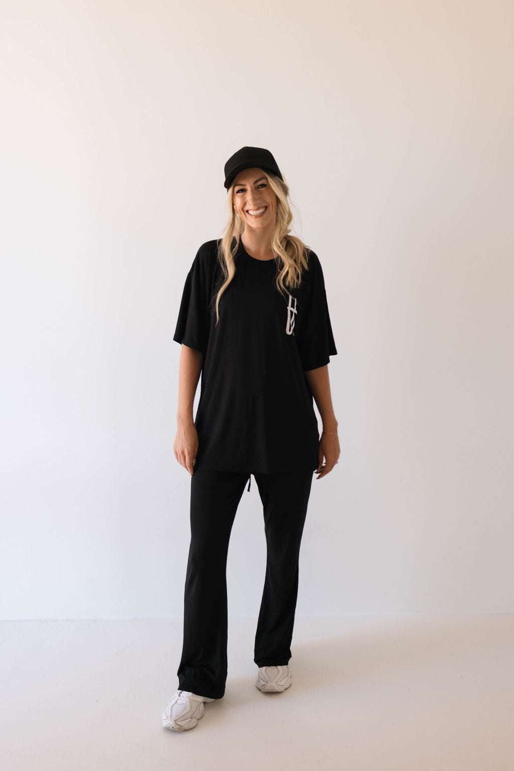 A person with long blonde hair is standing and smiling against a plain background. They are wearing a black cap, black pants, white sneakers, and a stylish yet comfortable Short Sleeve Women's Bamboo Lounge Set from forever french baby. Their relaxed attire and cheerful expression give off a casual, comfortable vibe.