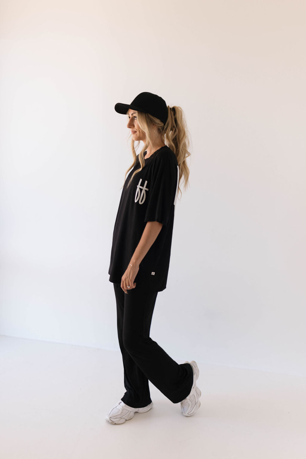 A person wearing a black outfit from forever french baby's Short Sleeve Women's Bamboo Lounge Set in Black ff Signature, consisting of a breathable bamboo fabric T-shirt, pants, and a cap, stands against a plain white background. They have long blonde hair tied back in a ponytail and are wearing white sneakers. The T-shirt features a small white logo on the front.