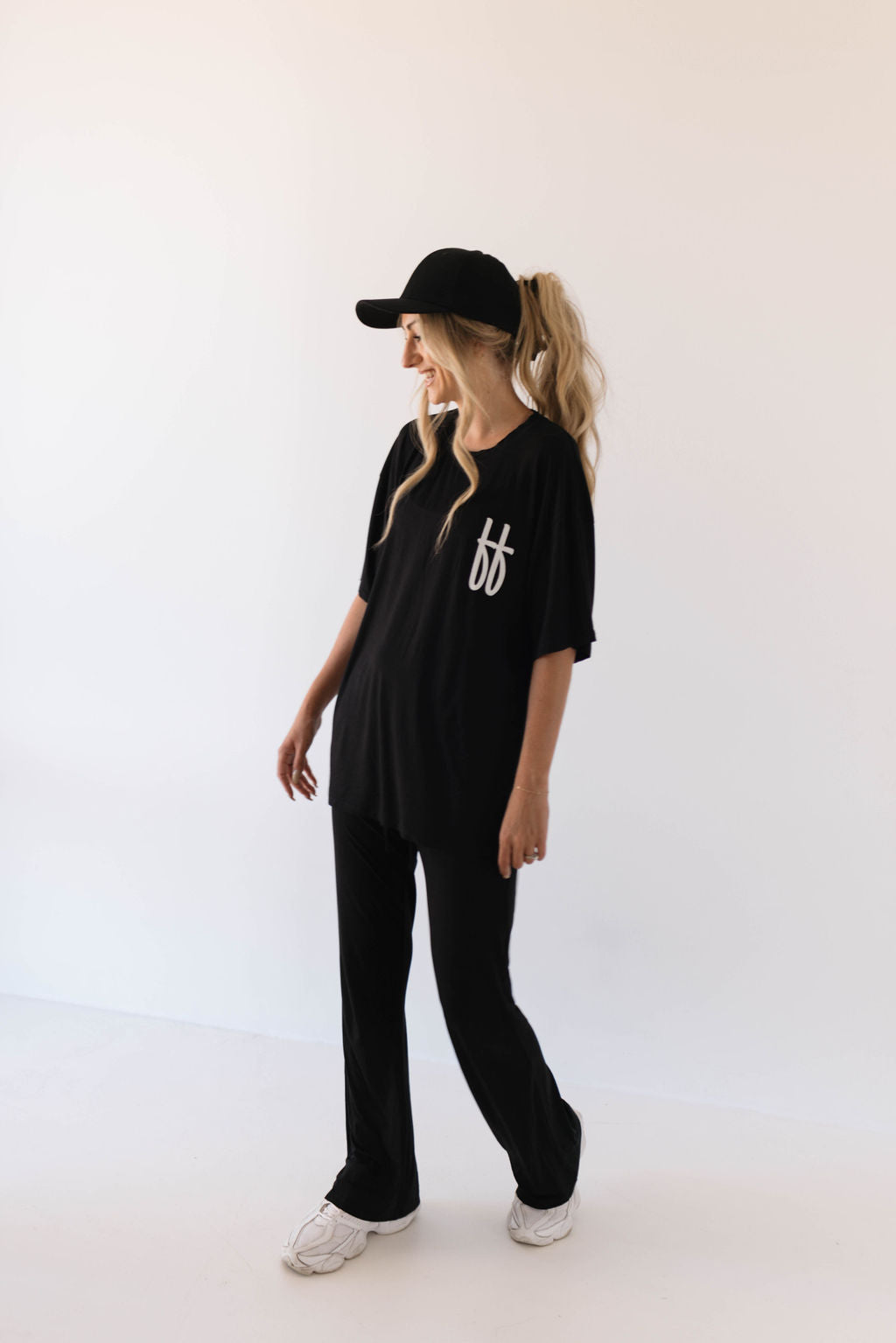 A woman stands against a simple white background wearing the "Short Sleeve Women's Bamboo Lounge Set" in black by forever french baby, featuring an oversized black t-shirt with the white "ff" signature on the chest and matching black pants. She has long blonde hair tied back and is looking to the side while smiling. She completes her outfit with white sneakers made of breathable bamboo fabric.