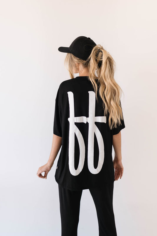 A person with long, blonde hair in a ponytail wears a black baseball cap and an oversized Short Sleeve Women's Bamboo Lounge Set | Black ff Signature by forever french baby with large white stylized letters on the back. They are also wearing black pants and are facing away from the camera against a plain white background.