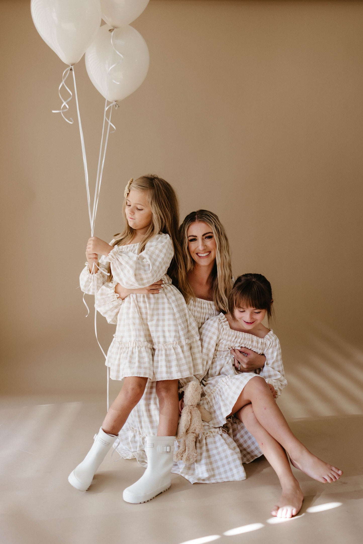 A woman and two young girls sit on the floor against a beige background, all wearing matching Women's Dresses in Gingham print by forever french baby. One of the young girls holds white balloons, while the other hugs a stuffed toy. They all look happy and relaxed.