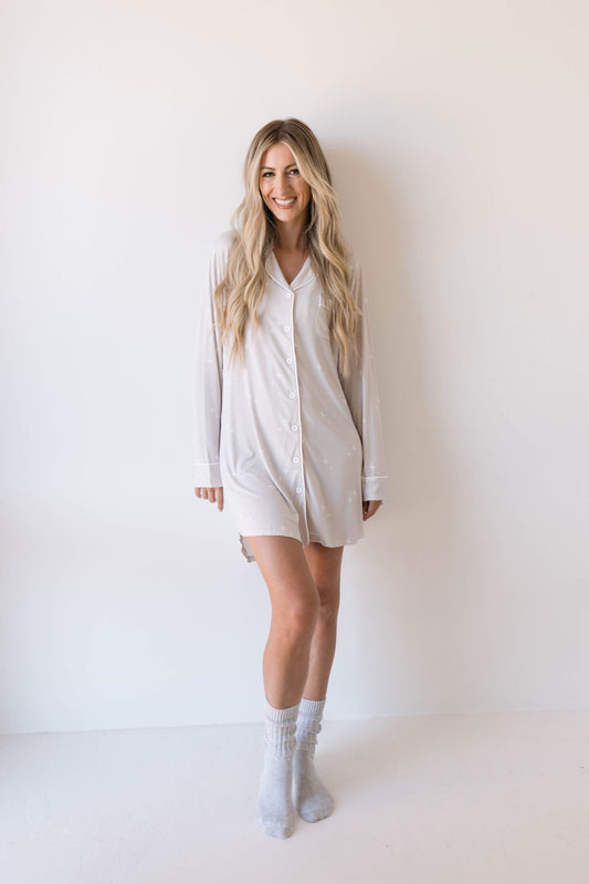 A woman with long blonde hair stands against a plain white wall, smiling in a light-colored, long-sleeve Women's Bamboo Sleeping Dress from forever french baby that extends to her mid-thigh. She is wearing white socks and has her feet slightly apart. This eco-friendly sleeping dress, named Sparkle!, offers hypo-allergenic comfort for peaceful nights.