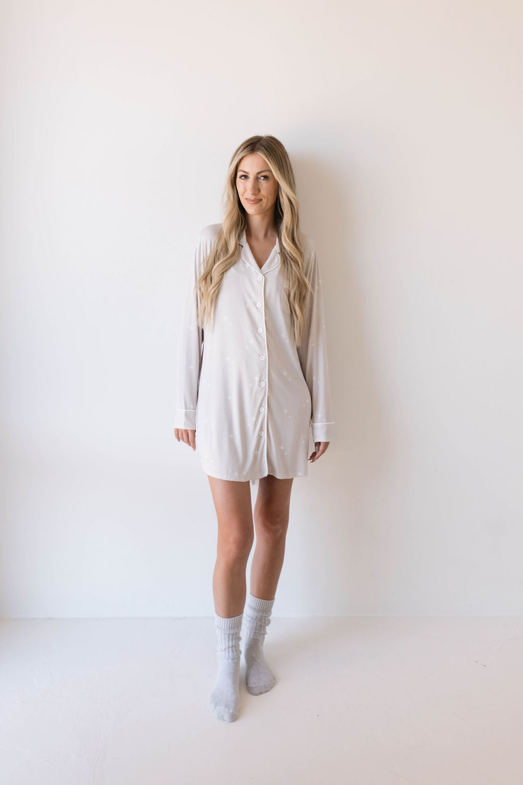 A woman with long blonde hair is standing against a plain white background. She is wearing an eco-friendly, light-colored, oversized button-up shirt from forever french baby’s Women’s Bamboo Sleeping Dress line—called Sparkle!—and light grey crew socks. She is looking at the camera and smiling slightly.