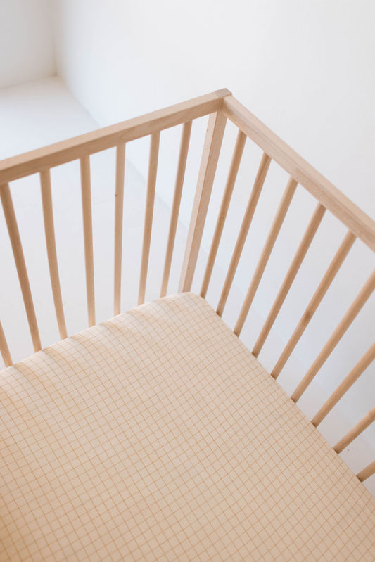 Close-up of a wooden crib with light brown vertical slats and a beige mattress featuring the Muslin Crib Sheet | Golden Grid by forever french baby. The muslin cotton sheet adds a soft touch in the bright, minimalist room with white walls.