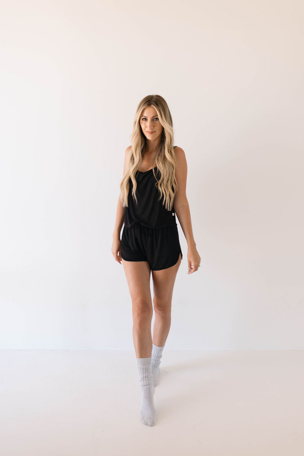 A woman with long blonde hair is standing against a plain white background. She is wearing the Cami Women's Bamboo Set in Midnight from forever french baby and light gray socks. Looking directly at the camera with a neutral expression, her right leg is slightly forward as if walking.