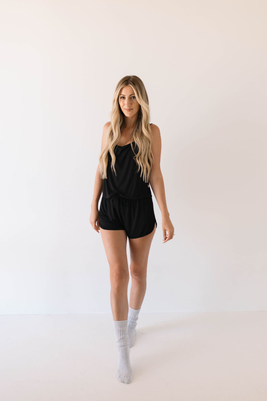 A woman with long blonde hair is walking toward the camera against a white background. She is wearing the Cami Women's Bamboo Set in Midnight from forever french baby, along with gray socks.