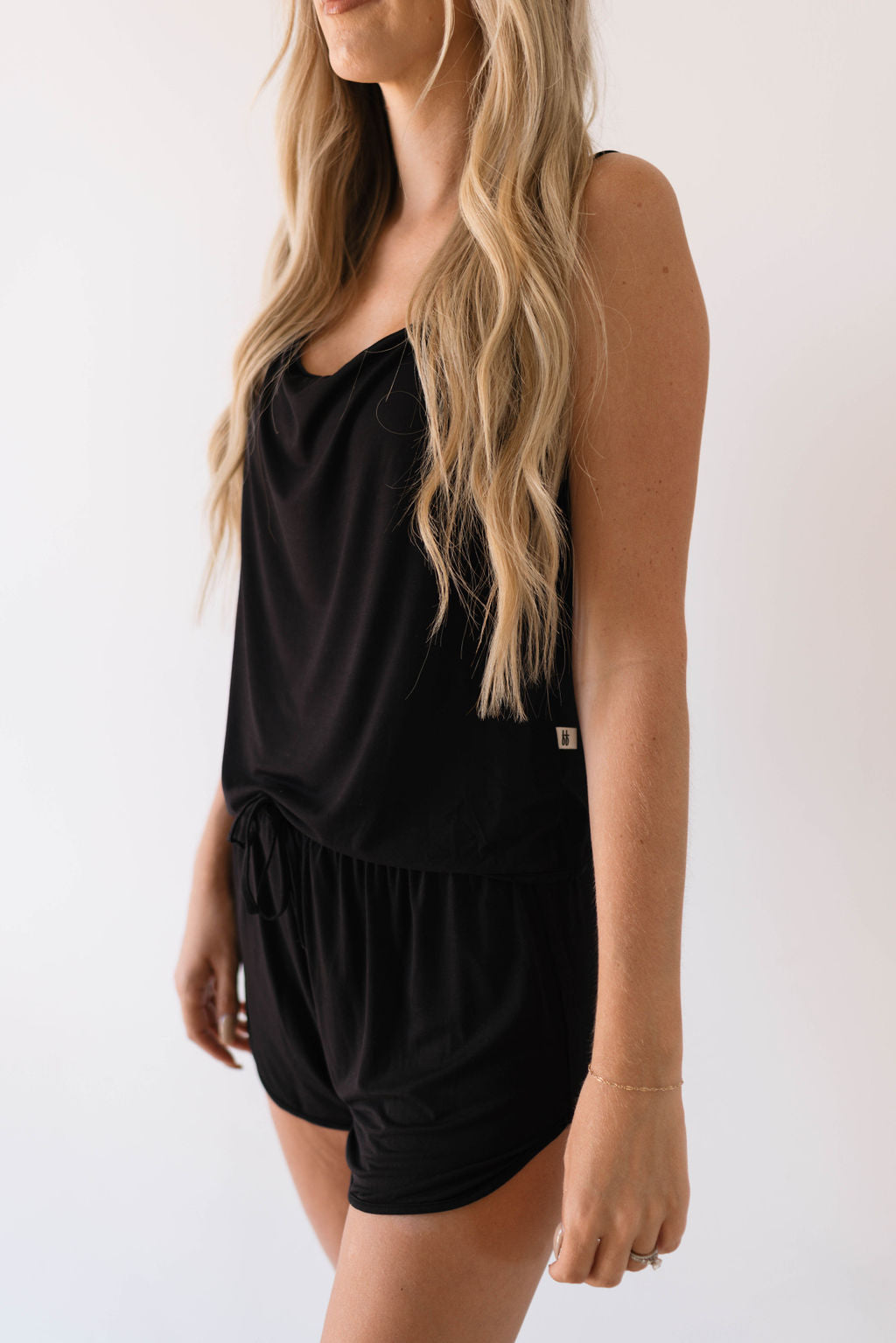 A person with long wavy hair, wearing the Cami Women's Bamboo Set in Midnight from forever french baby, stands against a plain white background. The black sleeveless top and matching black shorts are made from breathable bamboo fabric, and the outfit appears to be loose-fitting and comfortable. The person's head is turned slightly to the side.