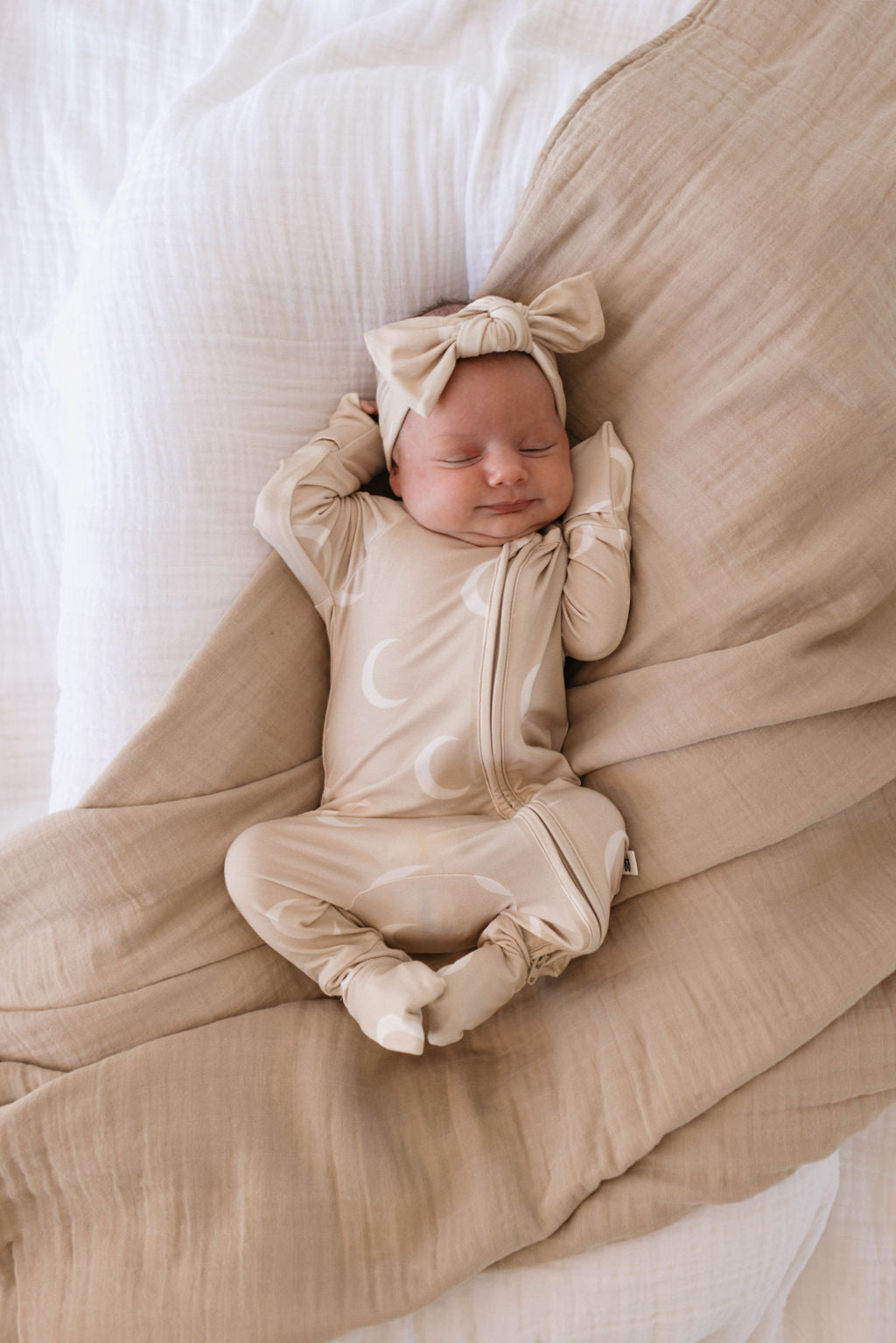 A baby is sleeping peacefully on a bed, dressed in a beige onesie with a crescent moon pattern. The baby is wearing a Bamboo Head Wrap | Luna from forever french baby and is surrounded by soft, beige and white bedding. The serene expression and arms raised above the head complete this precious moment.