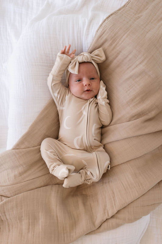 A baby lies on a bed, wrapped in a light beige blanket. The baby is wearing a cream-colored onesie with moon patterns and a Bamboo Head Wrap | Luna from forever french baby, complete with a bow. The baby has one arm raised, showcasing the elegance of babies and toddlers head wraps.