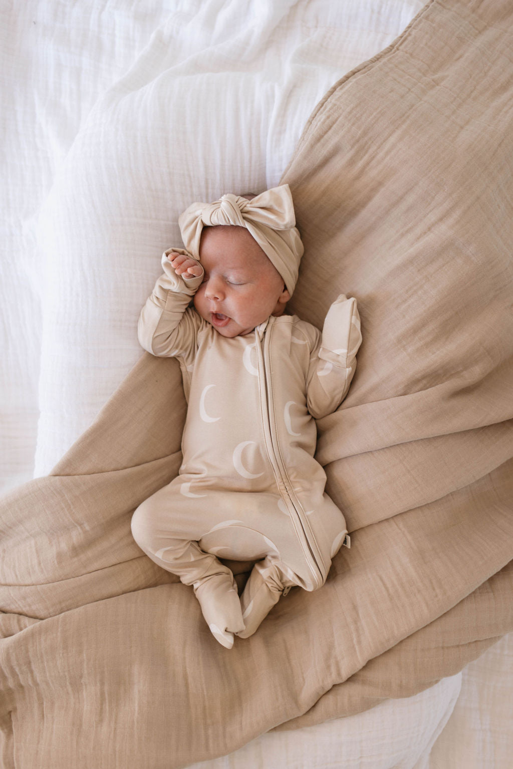 A baby sleeps on a bed, swaddled in a beige blanket. The baby wears a matching beige onesie with a moon design and a Forever French Baby Bamboo Head Wrap | Luna with a bow. The bed has white and beige linens. The baby’s hand is near their face while they rest peacefully.