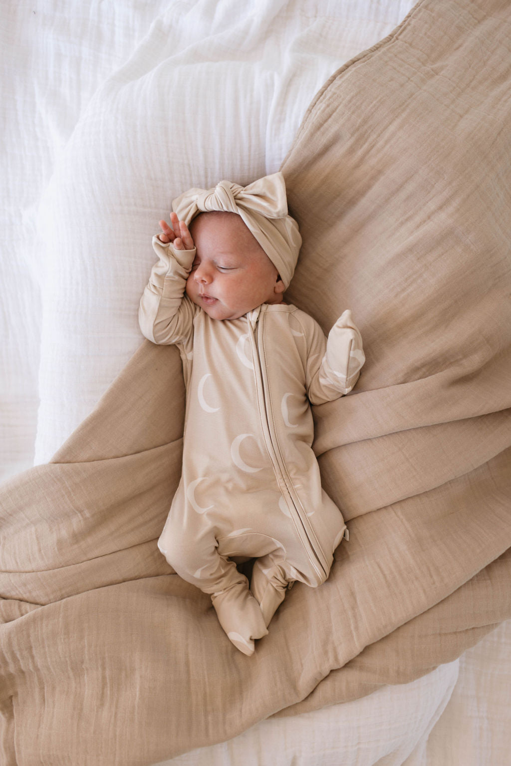 A sleeping newborn baby is dressed in a soft beige onesie and a matching "Bamboo Head Wrap | Luna" by forever french baby, adorned with a bow. The baby is lying on a white bed, partially covered with a beige blanket. The serene atmosphere suggests a peaceful, comfortable rest.