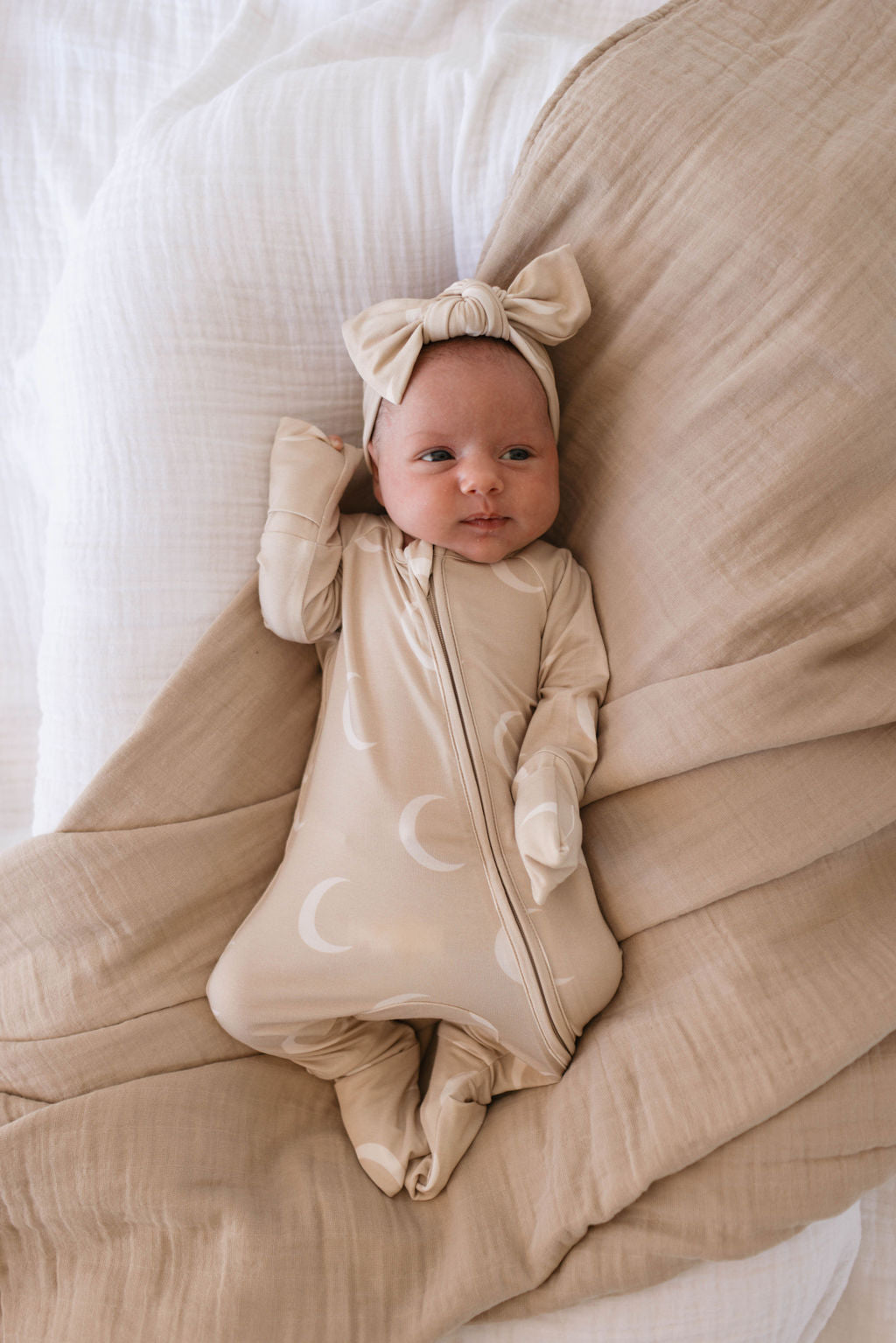 A baby lies on a white and beige blanket, dressed in a beige onesie with white crescent moon patterns. The baby also wears the "Bamboo Head Wrap | Luna" from forever french baby, and their small hand rests against the side of their head.