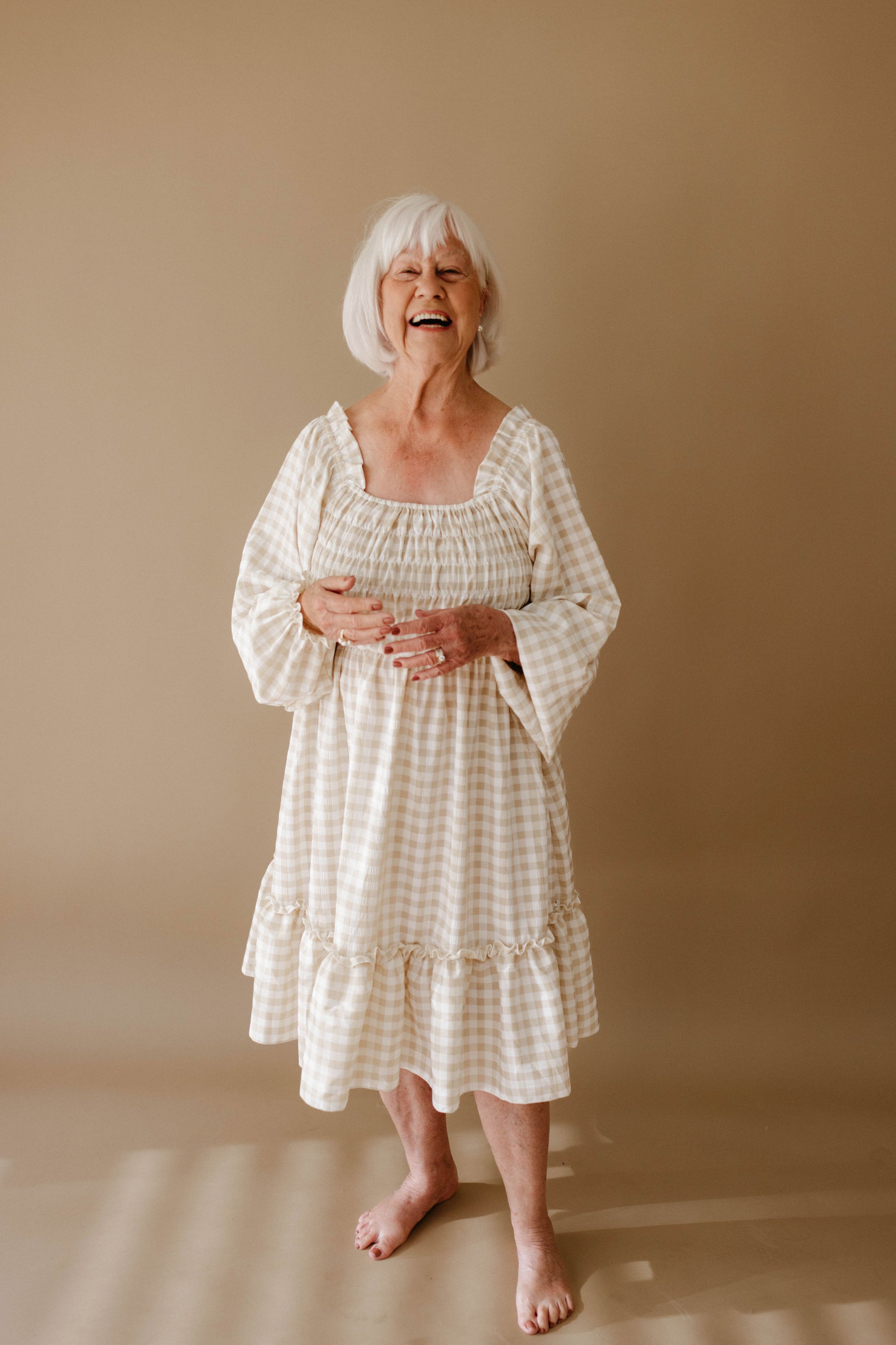 An elderly woman with short white hair stands barefoot, smiling in the Women's Dress | Gingham by forever french baby, against a plain beige background.