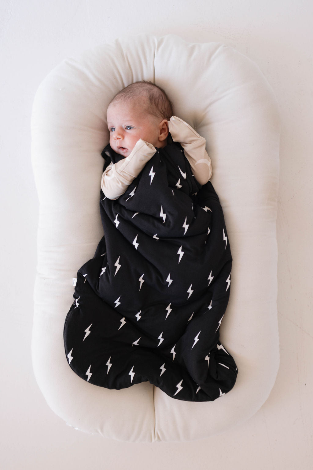 A baby is lying in a white cushioned crib, wrapped in the luxurious "Bamboo Sleep Sack | Midnight Lightning Bolt" from forever french baby. The baby's hands are tucked under their head, and they gaze off to the side with a calm expression.