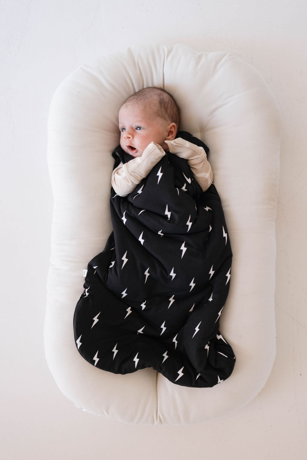 A baby wrapped in the luxurious Bamboo Sleep Sack | Midnight Lightning Bolt from forever french baby lies on a white cushioned surface. The baby, with light skin and short hair, gazes off to the side with a calm expression.