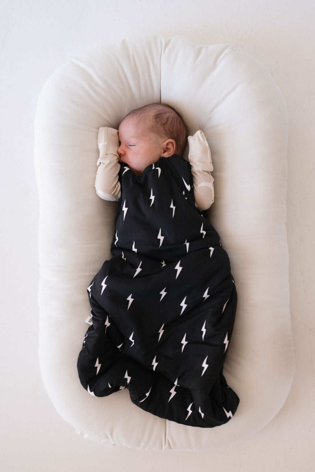 A baby sleeping on a cushioned surface, swaddled in the luxurious "Midnight Lightning Bolt" Bamboo Sleep Sack from Forever French Baby, adorned with white lightning bolt patterns. The baby is wearing white mittens and has its arms positioned near its head.