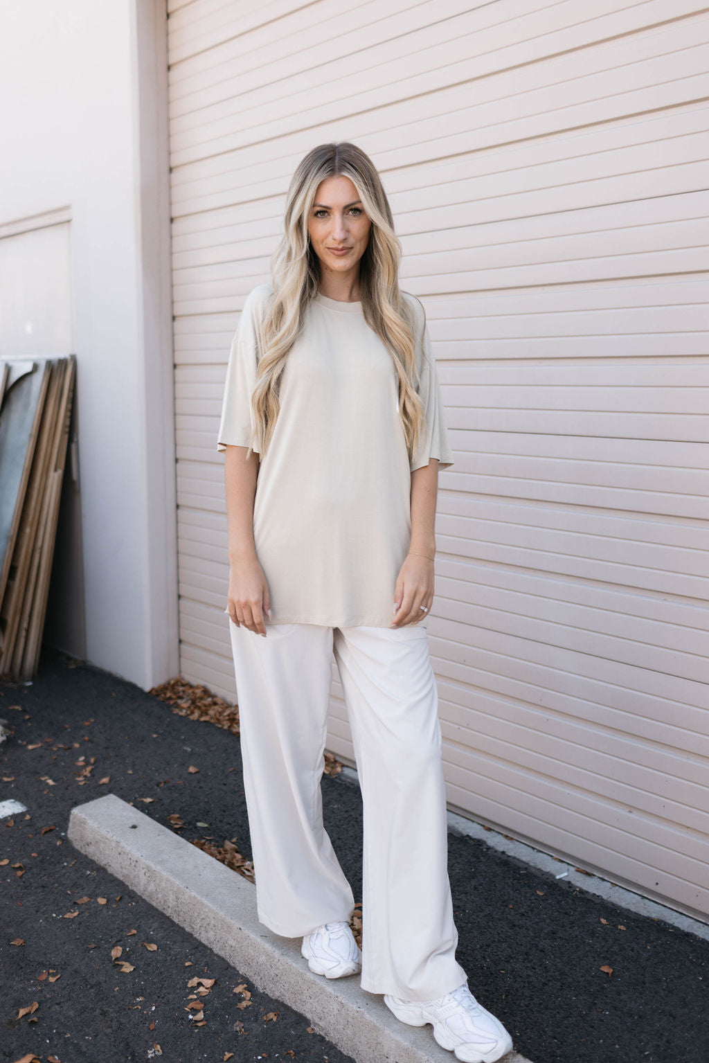 A woman with long blonde hair stands in front of a beige garage door, wearing an Adult Bamboo Tee in tan from forever french baby, paired with wide-legged white pants and white sneakers. She has a neutral expression, and her hands are relaxed by her sides, embodying hypoallergenic clothing comfort and style.