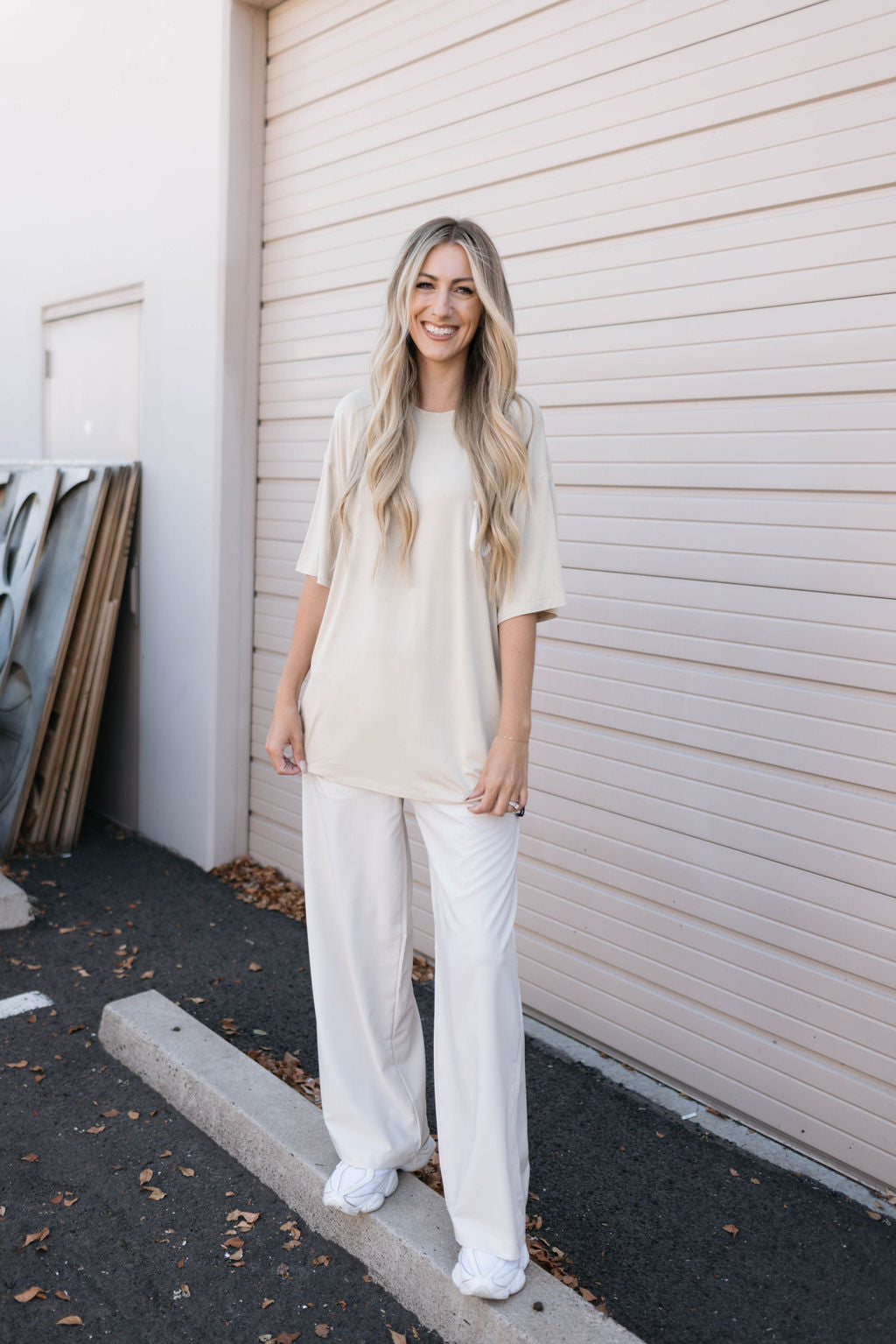 A person stands smiling in front of a closed garage door. They are wearing an oversized Adult Bamboo Tee in Tan from forever french baby, white loose-fitting pants, and white sneakers. They have long, wavy blonde hair and hold their hands casually in front of them. The background includes some stacked building materials and fallen leaves on the ground.