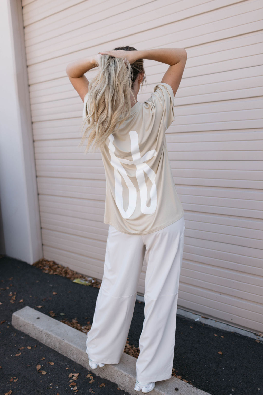 A person with long blonde hair stands with their back facing the camera, hands raised to tie their hair. They are wearing an Adult Bamboo Tee in Tan from forever french baby paired with loose-fitting white pants, standing in front of a beige garage door.