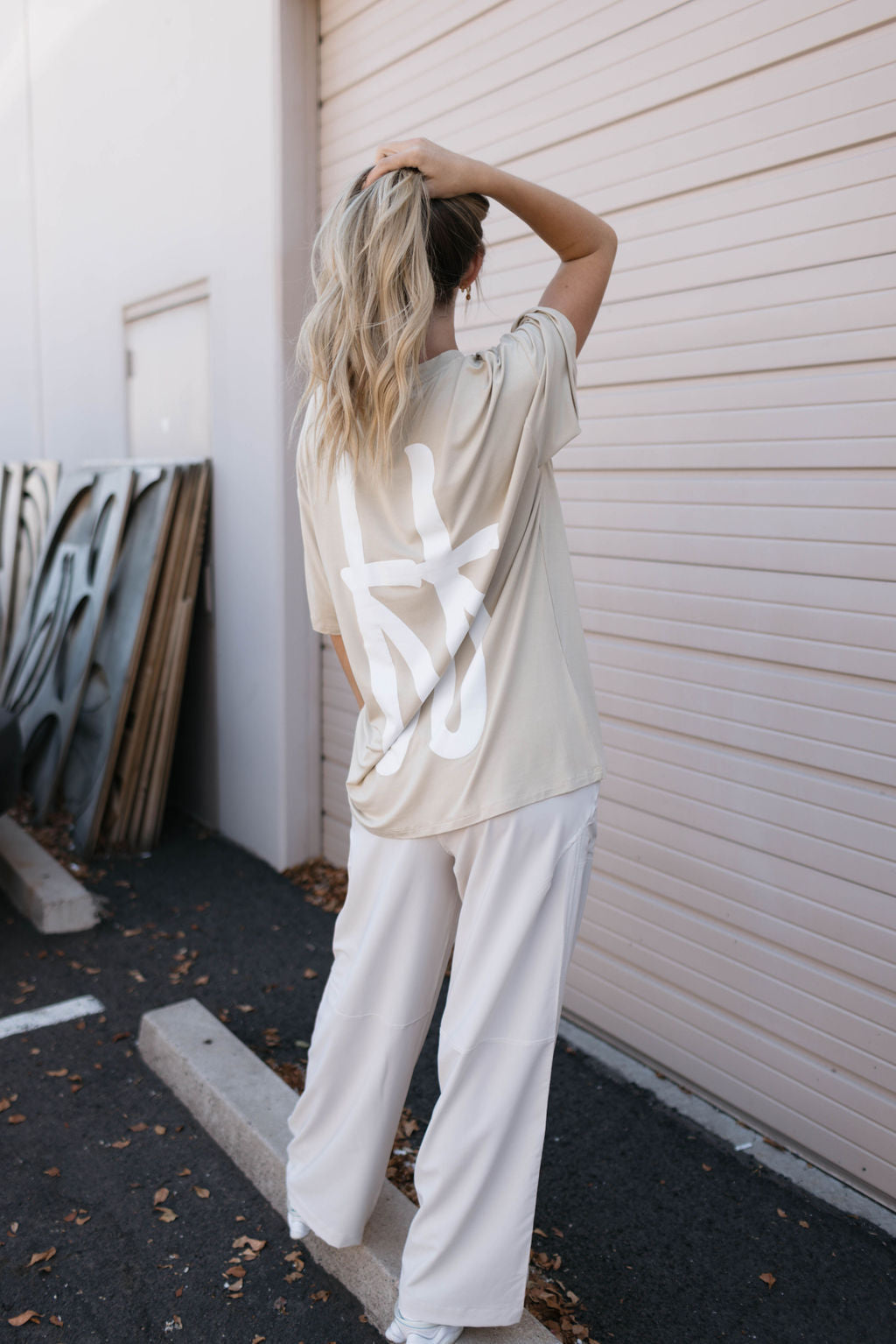 A person with long, blonde hair stands in front of a beige corrugated metal door, their hand resting on their head. They are wearing a tan oversized Adult Bamboo Tee from forever french baby and light-colored loose-fitting pants. Their face is not visible.