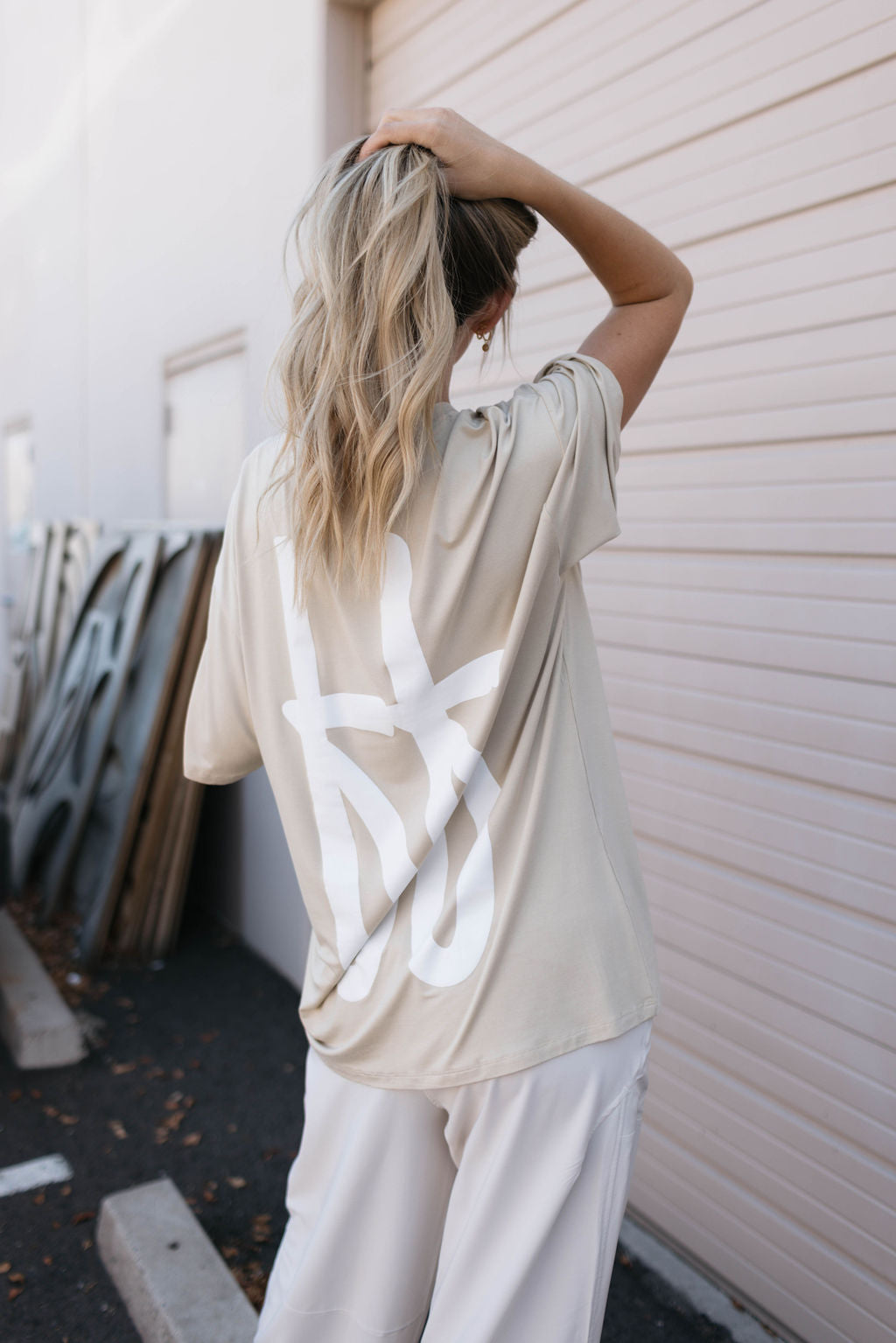 A person with long, wavy blonde hair, standing with their back to the camera and holding a ponytail, is wearing an oversized tan Adult Bamboo Tee from forever french baby. The tee features an intricate white graphic design on the back and they are also donning white pants. The scene is set against a beige garage door and stacked metallic panels in the background.