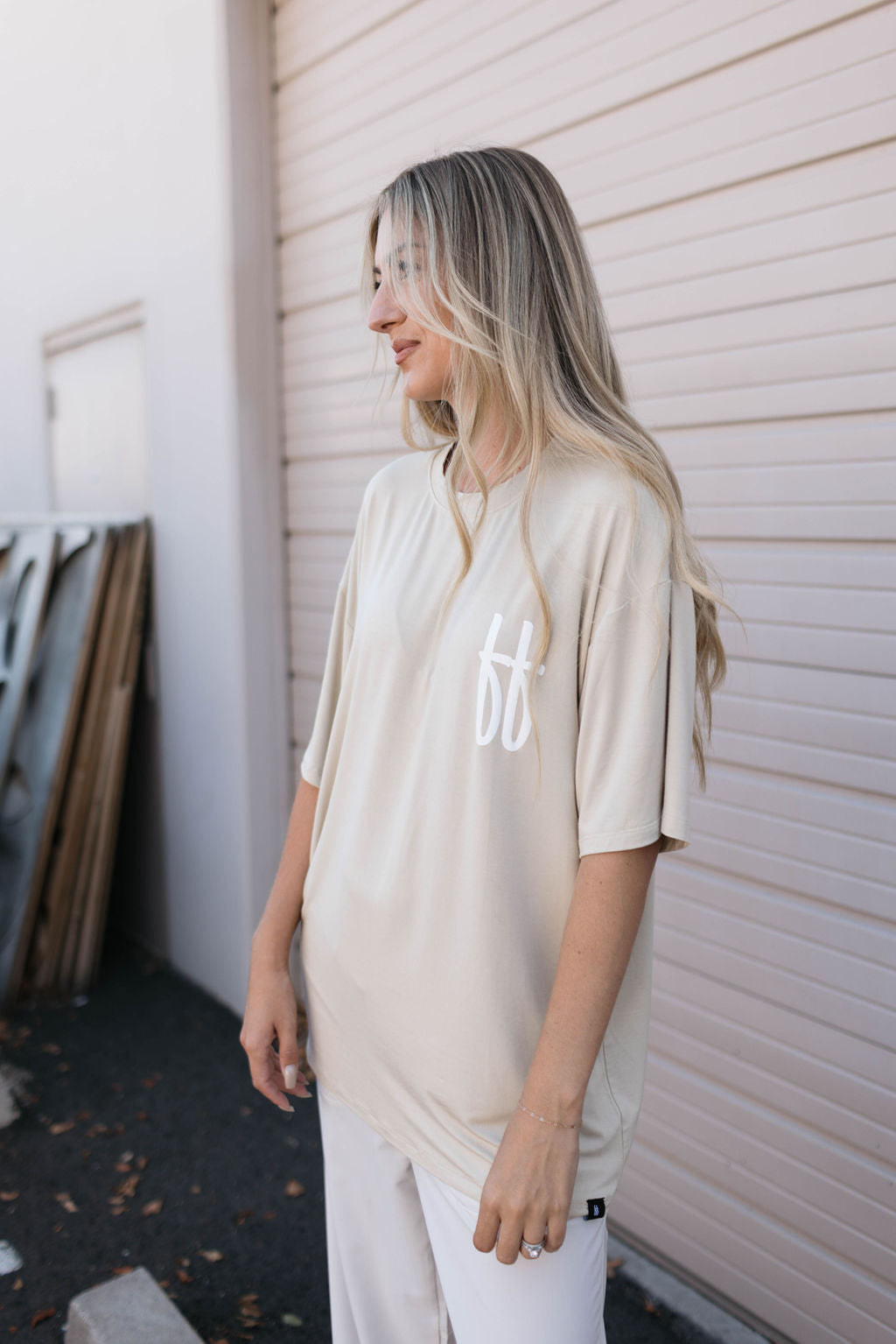 A person with long, light hair is standing outdoors, wearing an Adult Bamboo Tee in tan from forever french baby. The tee features a white logo on the left chest and is paired with white pants. The background showcases a light-colored, textured wall adorned with some wooden panels leaning against it.