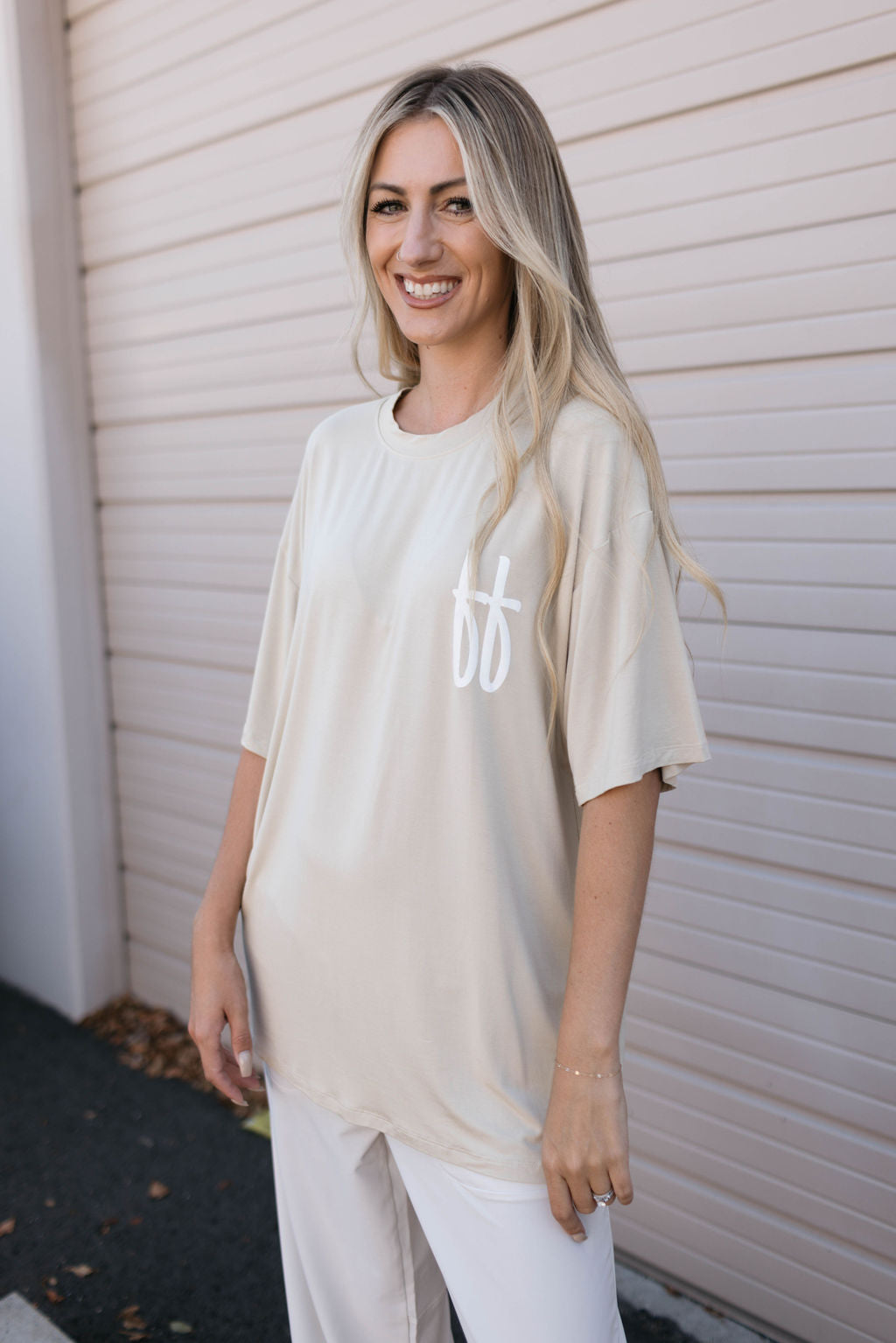 A woman with long blonde hair is smiling while standing in front of a light-colored garage door. She is wearing a beige Forever French Baby Adult Bamboo Tee in Tan ff, featuring a white stylized logo on the chest, paired with white pants.