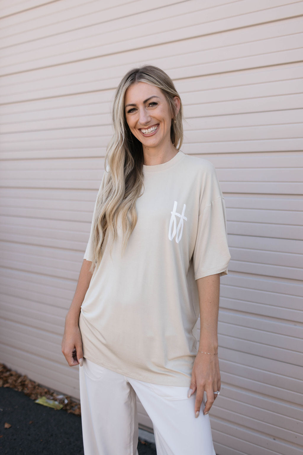 A smiling woman with long, wavy blonde hair stands in front of a beige garage door. She is wearing a tan oversized Adult Bamboo Tee from forever french baby with a white logo on the left side, paired with white pants. One hand is in her pocket while the other rests on her hip.
