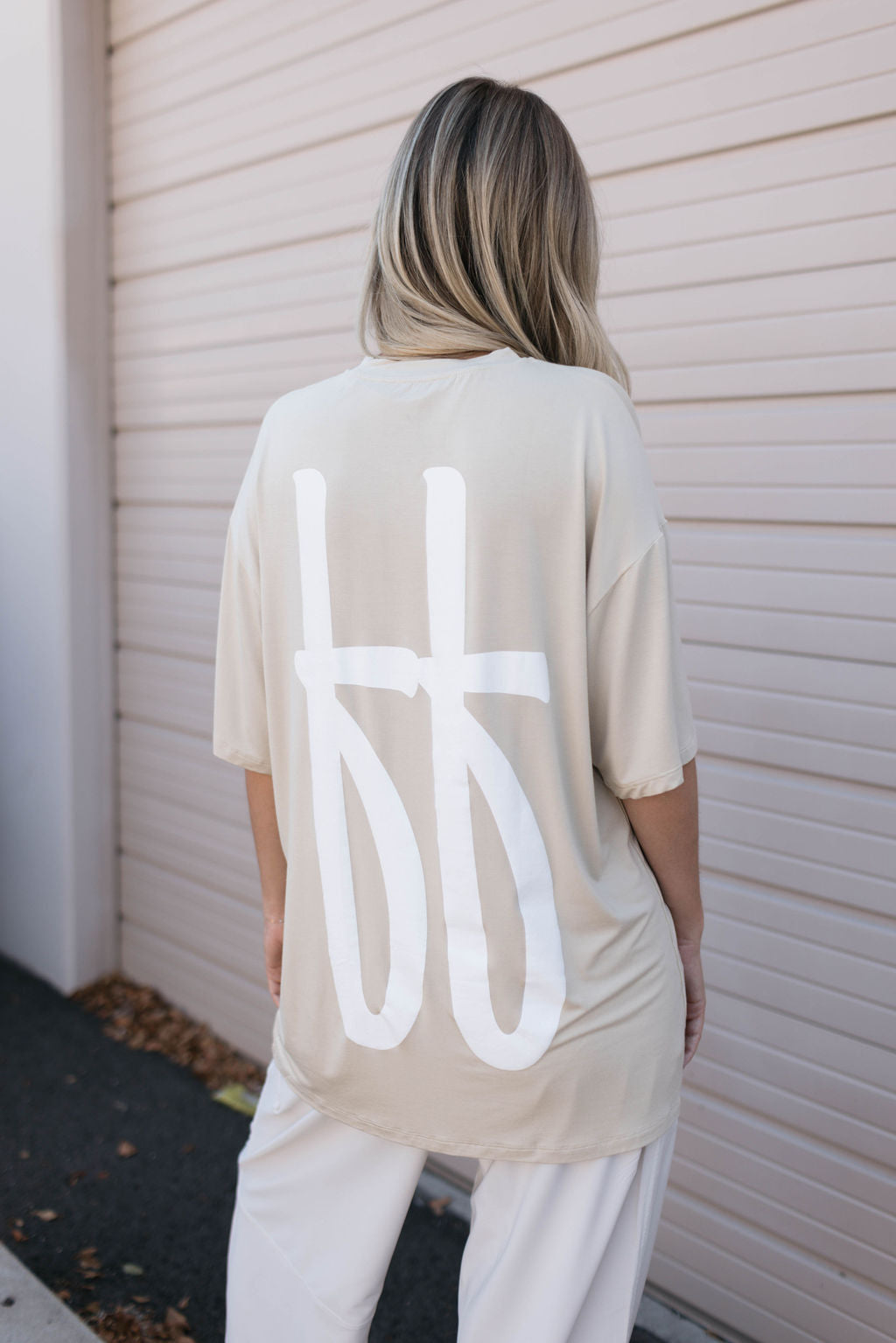 A person with long, blonde hair is facing away from the camera, wearing an oversized tan Adult Bamboo Tee from forever french baby. The beige tee features a large white graphic on the back. The person stands in front of a light-colored garage door, showcasing hypo-allergenic clothing that's stylish and comfortable.