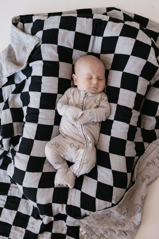 A serene infant in forever french baby's "Bamboo Zip Pajamas | Arm Day" rests peacefully on a soft, breathable black-and-white checkered blanket, with arms crossed over their chest.