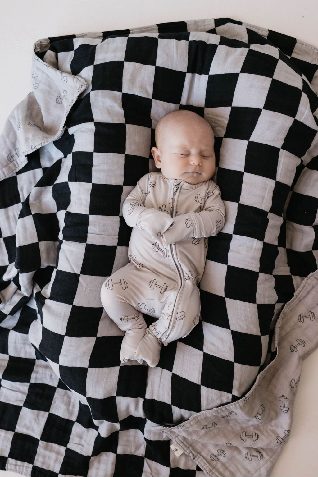 A baby sleeps peacefully on a large black and white checkered blanket, dressed in forever french baby's Bamboo Zip Pajamas | Arm Day onesie with small dumbbell patterns made from breathable fabric.