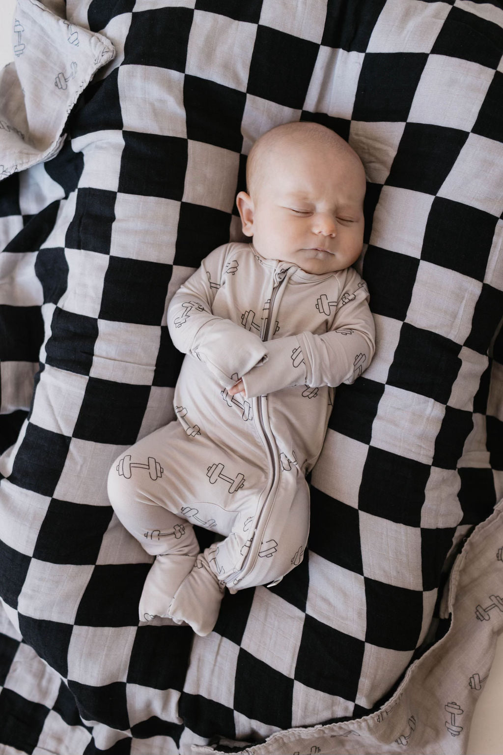 A sleeping baby in forever french baby's Bamboo Zip Pajamas | Arm Day rests peacefully on a black and white checkered blanket, wrapped in breathable fabric.