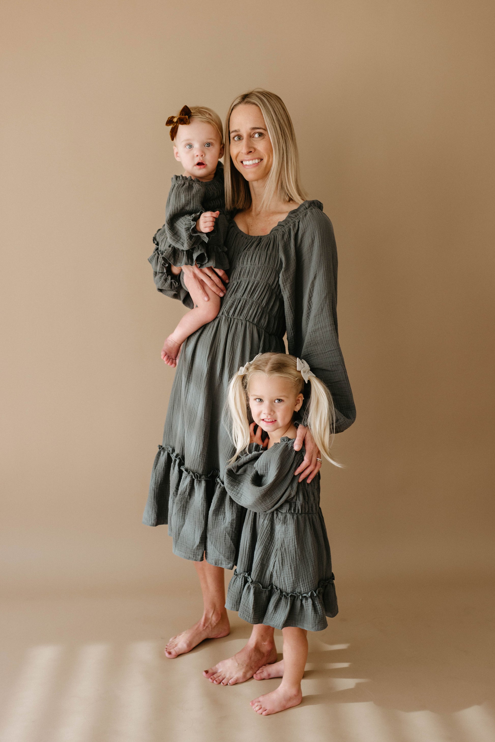 A woman, a toddler with a brown bow, and a blonde girl in pigtails stand barefoot on beige, all wearing matching forever french baby Meadow long-sleeve ruffled olive-green dresses.