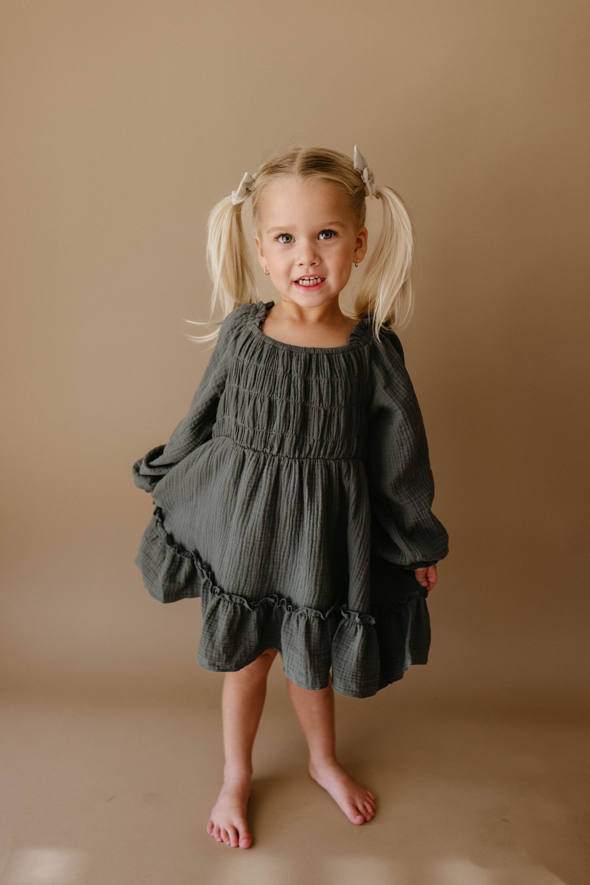 A smiling young girl with blonde pigtails wears the Meadow Child's Dress from forever french baby—a dark green, long-sleeved, ruffled dress—standing barefoot against a neutral background.