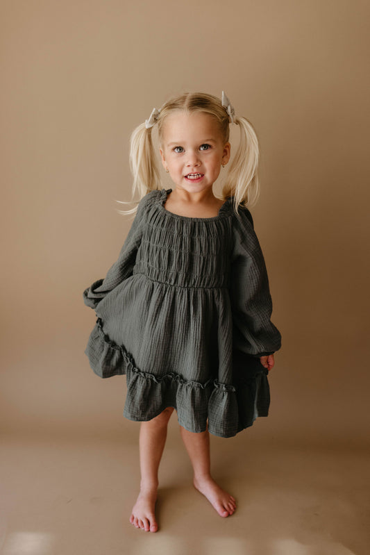 A smiling young girl with blonde pigtails wears the Meadow Child's Dress from forever french baby—a dark green, long-sleeved, ruffled dress—standing barefoot against a neutral background.