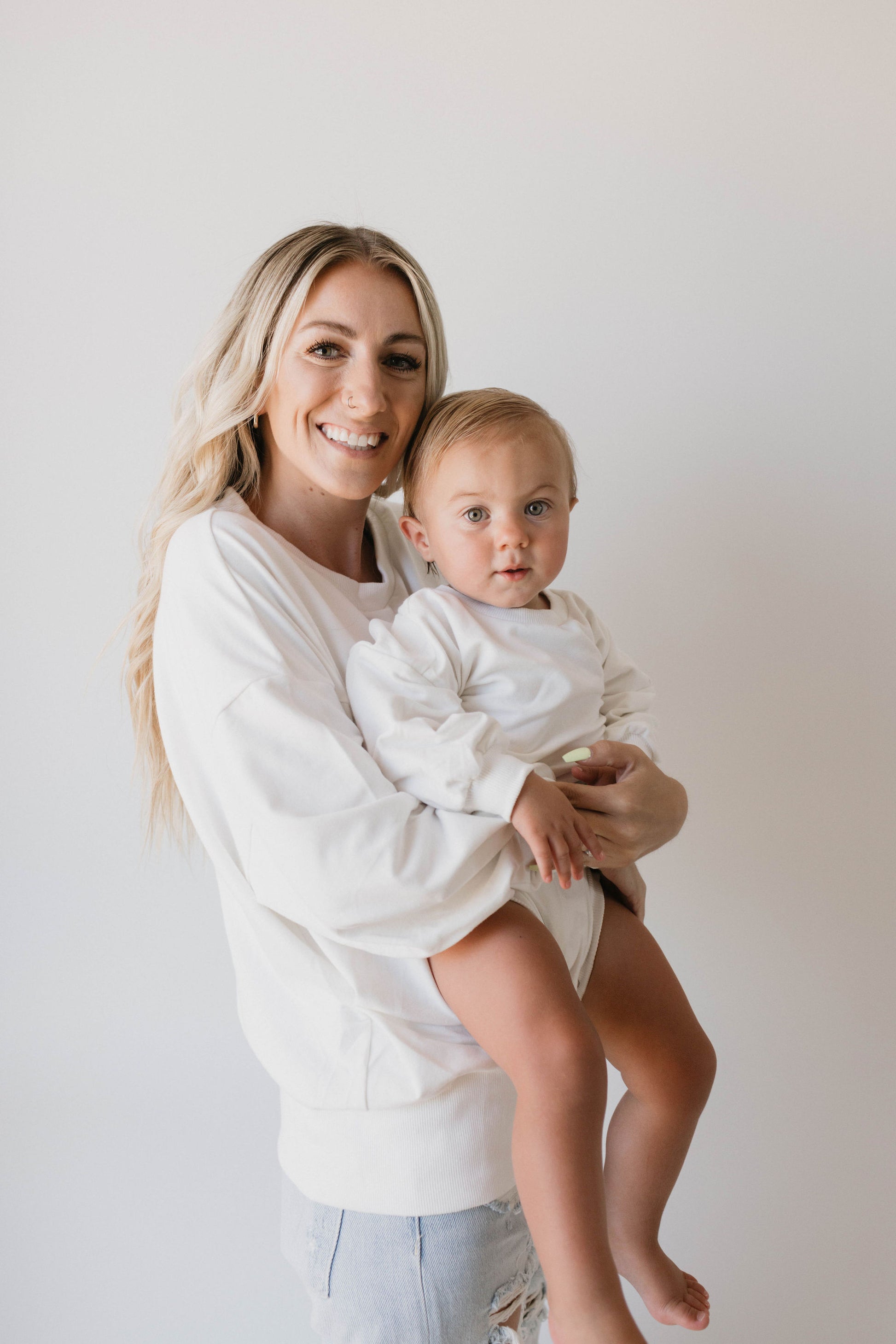 A woman with long blond hair, wearing a white sweater, smiles as she holds a baby dressed in the "Sweatshirt Romper | Dove" from forever french baby. They are standing against a plain white background.