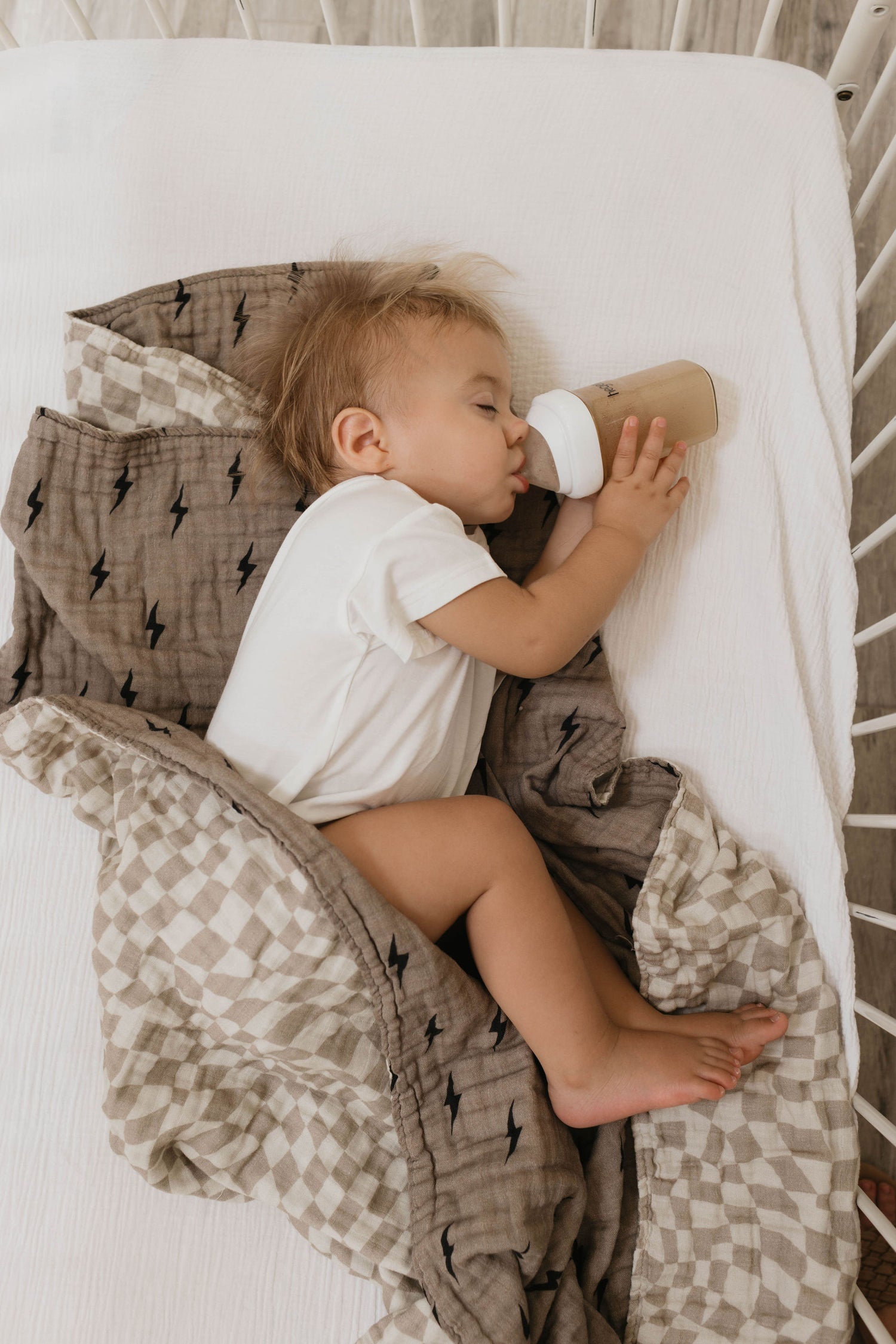 A toddler with light brown hair is lying in a crib with white bars, drinking from a baby bottle while holding it with both hands. The child is wearing a white onesie and is covered by the Muslin Quilt | Lightning Bolt & Wavy Checker from forever french baby, featuring a beige and grey lightning bolt pattern.